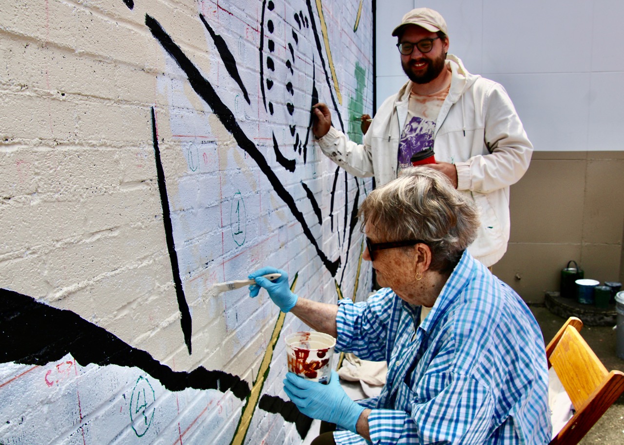 (Rick Miller/Olean Star) Mural designer and artist Alex Derrick watches Sunday as his grandmother, Sondra Fox, applies paint to “The Moment,” an Olean mural taking shape on the side of the Sports Locker open West State Street.