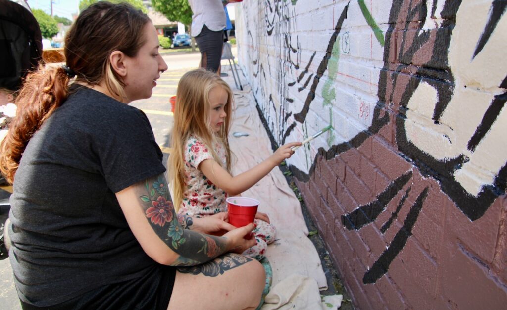 (Rick Miller/Olean Star) Sierra Churakos watches as her 4-year-old daughter Isla paints one the Sports Locker mural “The Moment” on Sunday.