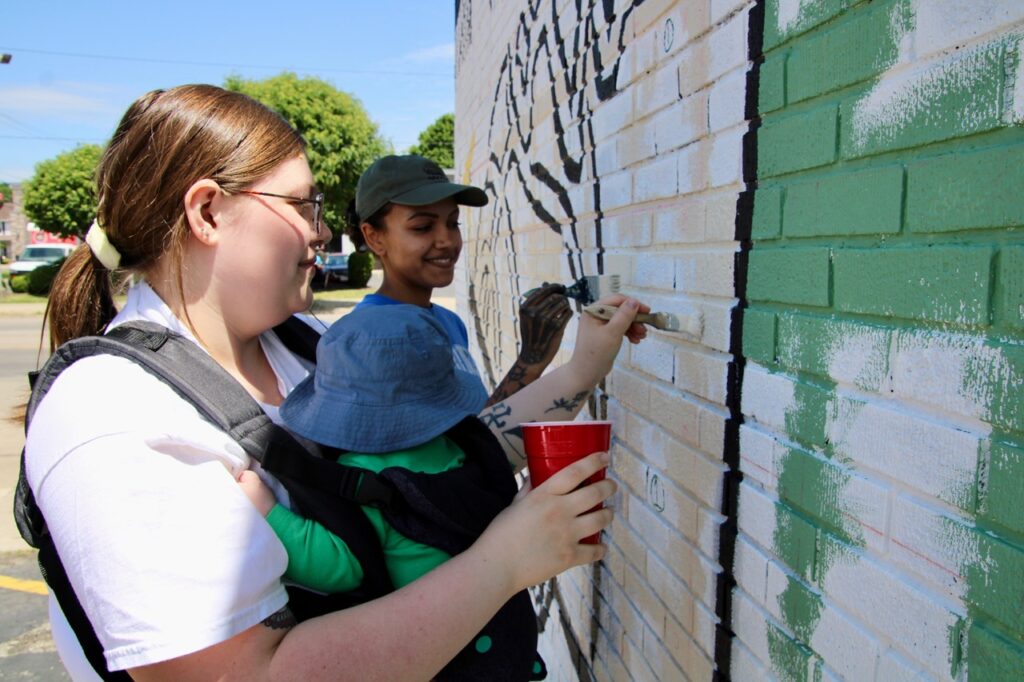 (Rick Miller/Olean Star) Cassidy Sawyer paints as her 4-month-old son, Oliver, tries to take a nap Sunday at the public painting of the mural “The Moment” on the side of the Sports Locker on West State Street. Own her left is Quinn Nova, an artist who has worked on other Olean murals.