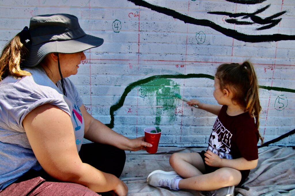 (Rick Miller/Olean Star) Hannah Derrick, sister of mural artist Alex Derrick, hold a cup of paint as Josephine paints a green color on “The Moment,” a West State Street mural at the Sports Locker.