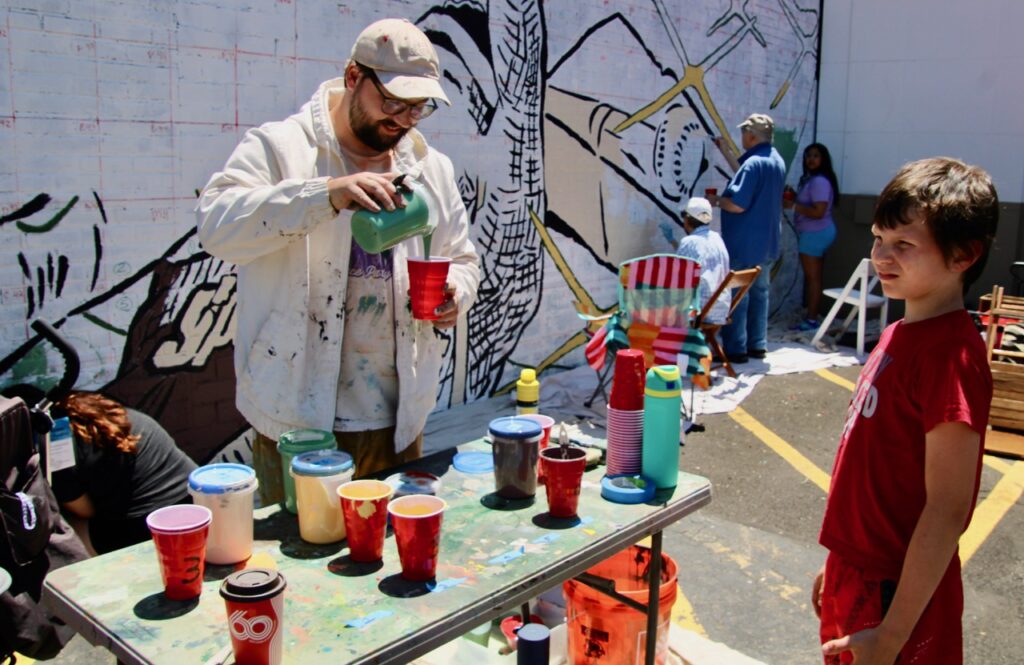 (Rick Miller/Olean Star) Mural designer and artist Alex Derrick mixes up a cup of paint Sunday for Matthew Barrick during the Public Paint Day for his mural “The Moment” on the Sports Locker building on West State Street.