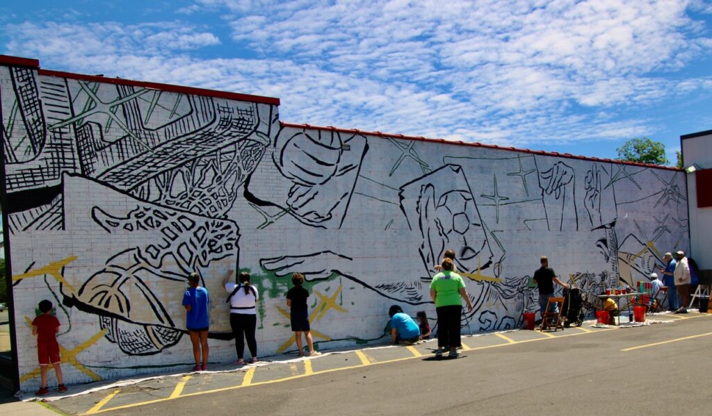 (Rick Miller/Olean Star) Members of the public grabbed paint brushes to participate in a Public Paint Day Sunday to work on Olean’s latest public art, a mural called “The Moment” on the side off the Sports Locker building on West State Street. Designer and artist Alex Derrick and others hope to finish the mural in the coming week.