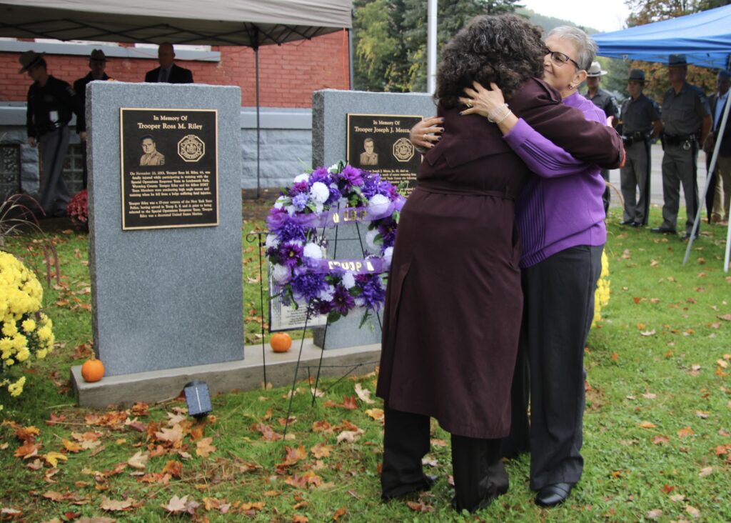 (Rick Miller/Olean Star)  Ret. Sgt Heidi Riley and Linda Mecca, widows of Troopers Ross M. Riley and Joseph J. Mecca Jr., both of whom died in the line of duty, hug after unveiling memorials outside the Olean State Police Substation Sunday.
